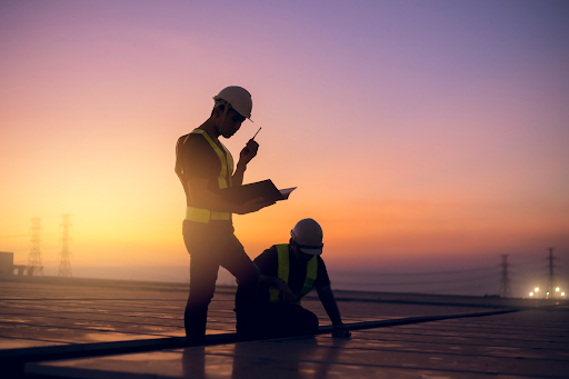 Two professional roofers are inspecting a large commercial roof at sunrise. One is holding a walkie talkie.